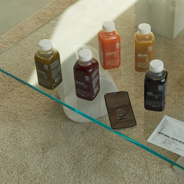 Fünf bunte Saftflaschen, ein Smartphone und eine Packung Gesichtsmasken liegen auf einem Glastisch über einem beigen Teppich. Die Gegenstände suggerieren einen Fokus auf Gesundheit, Wellness und Selbstfürsorge.