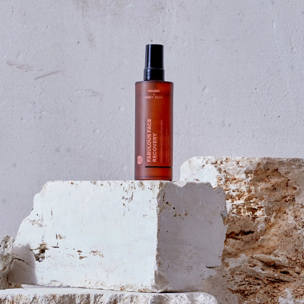 Eine braune durchsichtige Sprühflasche mit der Aufschrift Fabulous Age Recovery steht aufrecht auf strukturierten weißen Steinblöcken vor einer hellgrau verputzten Wand.