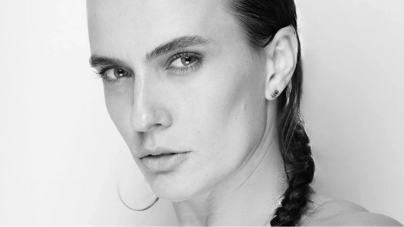 Eine Person mit heller Haut und geflochtenem dunklem Haar blickt mit ernster Miene direkt in die Kamera und trägt einen kleinen Ohrstecker vor einem schlichten hellen Hintergrund.