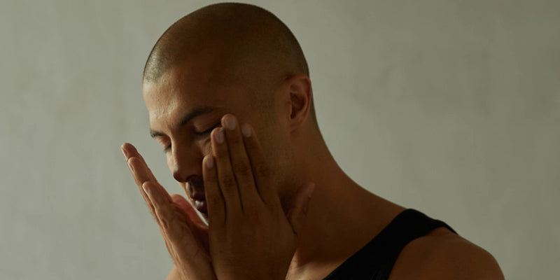 Eine Person mit rasiertem Kopf, die ein schwarzes Tank-Top trägt, berührt mit beiden Händen ihr Gesicht, die Augen geschlossen, vor einem schlichten, hellen Hintergrund.