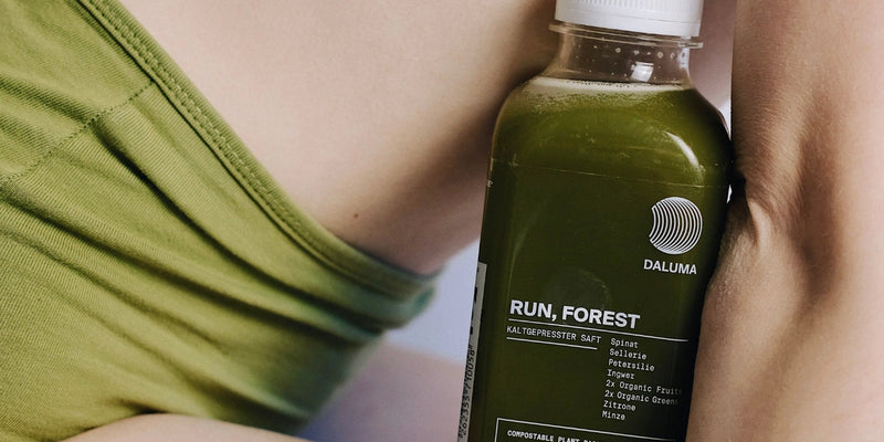 Eine grüne Saftflasche mit der Aufschrift RUN, FOREST von DALUMA wird dicht an das nackte Bein einer Person mit grüner Kleidung gehalten. Die Flasche enthält kaltgepressten Saft mit Grünkohl, Petersilie und anderen Zutaten.