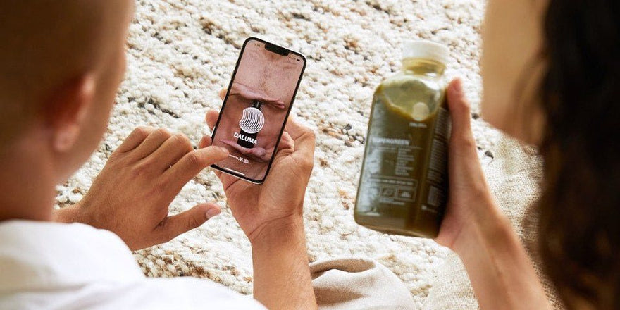 Person hält ein Smartphone mit geöffneter DALUMA App, daneben hält eine zweite Person eine Flasche Supergreen Saft über einem hellen Wollteppich