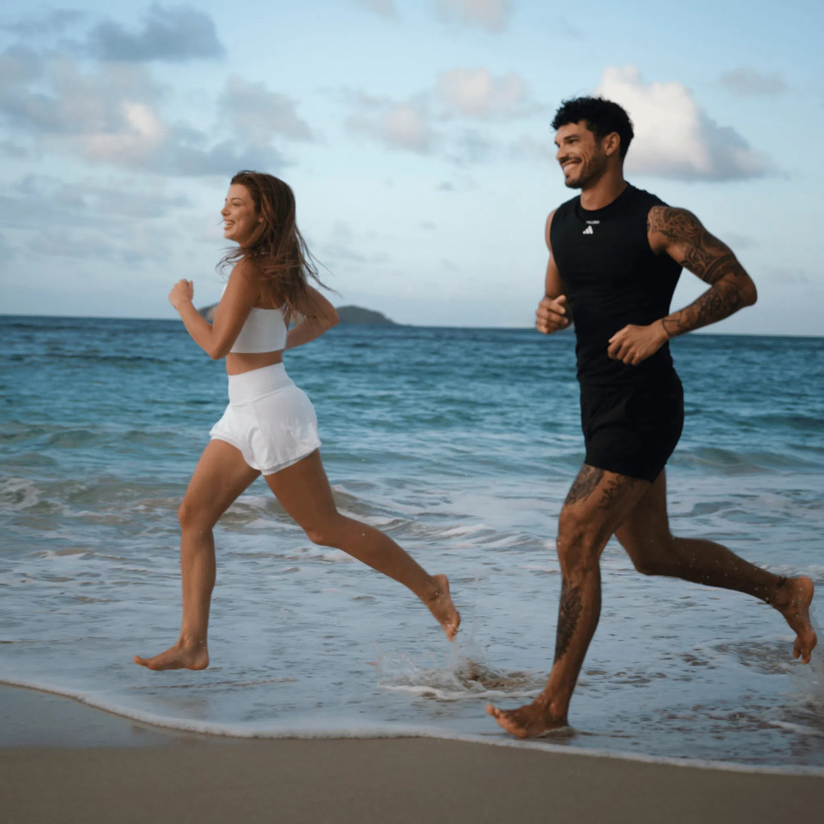 Lachende Frau in weißem Sportoutfit und Mann in schwarzem Sportoutfit joggen barfuß am Strand durch die Brandung, im Hintergrund Meer und eine kleine Insel