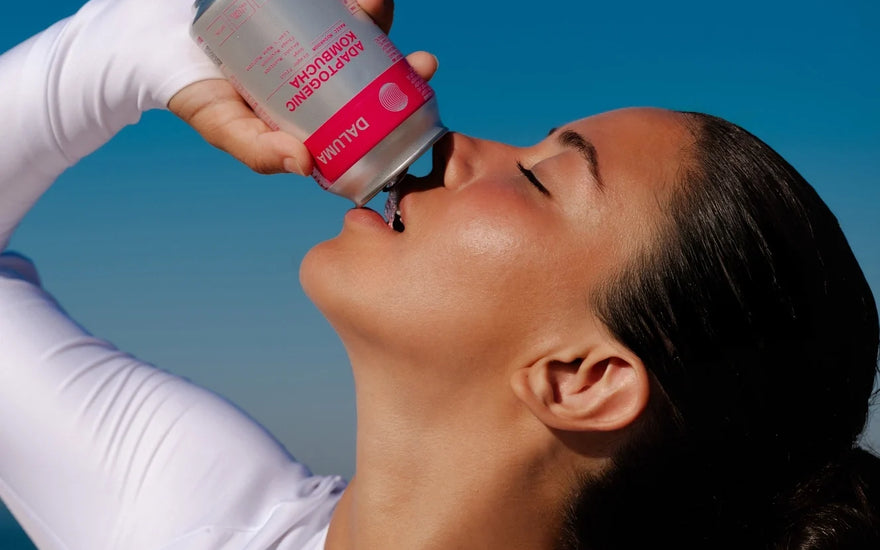 Frau im weißen Sportoutfit trinkt mit geschlossenen Augen aus einer Dose DALUMA Adaptogenic Kombucha vor strahlend blauem Himmel