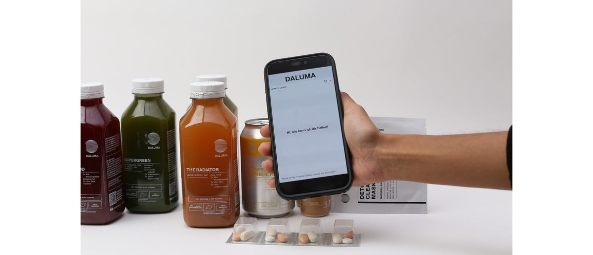 Smartphone mit dem DALUMA Health Coach Chat vor kaltgepressten Säften, Kombucha und Detox Clearing Mask.