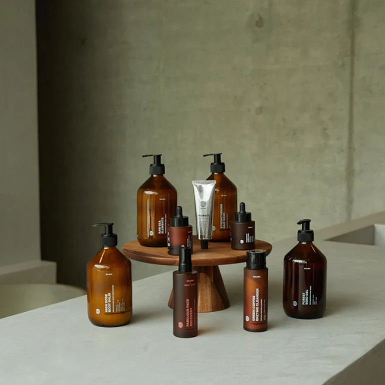 Eine Reihe von bernsteinfarbenen und braunen Hautpflegeflaschen und -tuben, die auf einem kleinen Holzständer vor einem minimalistischen Betonhintergrund angeordnet sind.