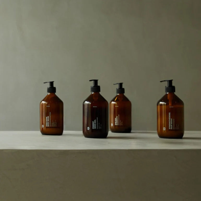 Vier braune Pumpflaschen mit minimalen weißen Textetiketten sind auf einer hellgrauen Fläche vor einem neutralen grauen Hintergrund angeordnet. Die Szene ist einfach und stilvoll und erinnert an eine moderne, minimalistische Ästhetik.