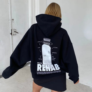Eine Person mit hellbraunem Haar trägt einen übergroßen schwarzen Kapuzenpullover mit einer weißen Flaschengrafik und dem Wort REHAB auf dem Rücken und steht in einem Innenraum in der Nähe einer weißen Wand und Tür.