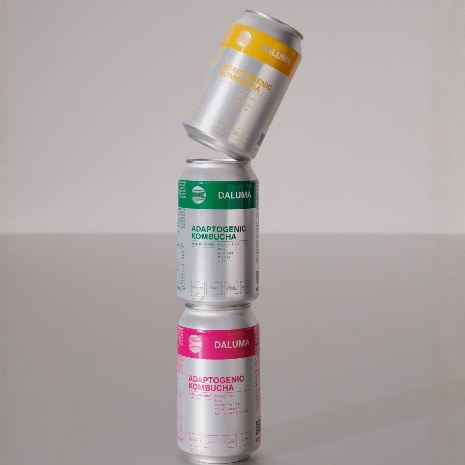 Drei Dosen Daluma Adaptogenic Kombucha sind auf einer spiegelnden Oberfläche gestapelt, mit gelben, grünen und rosafarbenen Etiketten, und die oberste Dose ist schräg gestellt.