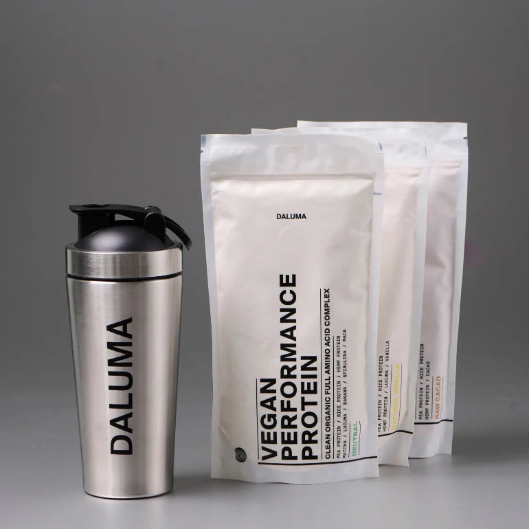 Ein Edelstahl-Shaker mit der Aufschrift DALUMA steht neben drei weißen Beuteln mit DALUMA Vegan Performance Protein vor einem schlichten grauen Hintergrund.