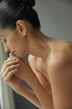 Eine Frau mit dunklem Haar in einem Dutt sitzt oben ohne, die Augen geschlossen, und berührt sanft ihre Lippen mit den Händen in weichem natürlichem Licht, was eine kontemplative und ruhige Stimmung erzeugt.