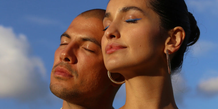 Ein Mann und eine Frau mit geschlossenen Augen sonnen sich im Sonnenlicht vor einem blauen Himmel mit Wolken. Die Frau trägt kräftigen blauen Eyeliner und große Ohrringe, und beide haben einen heiteren Gesichtsausdruck.