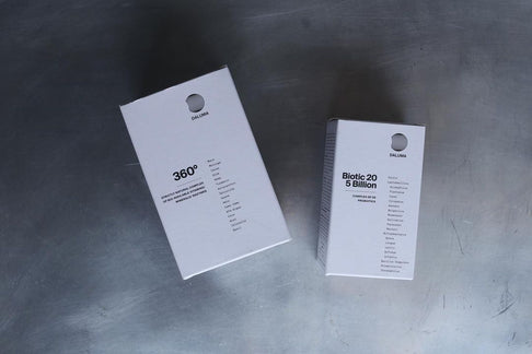 Zwei weiße rechteckige DALUMA-Ergänzungsdosen, darunter 360 DAILY ESSENTIALS SUPPLEMENT, ruhen auf einer Metalloberfläche. Beide haben ein minimalistisches Design, ein rundes Logo, Inhaltsstofflisten und heben natürliche Inhaltsstoffe hervor.