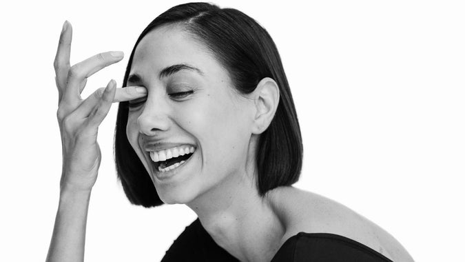 Eine Frau mit kurzen dunklen Haaren und einem schwarzen Oberteil lächelt breit und lacht, wobei sie mit der linken Hand ihr Gesicht in der Nähe der Nase berührt. Der Hintergrund ist einfarbig weiß.