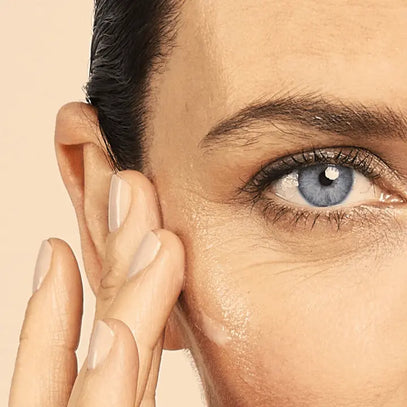 Nahaufnahme einer Person mit heller Haut und blauen Augen, die mit ihren Fingern sanft die Haut in der Nähe ihres rechten Auges eincremt.