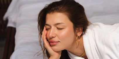 Eine Frau mit geschlossenen Augen und einem entspannten Gesichtsausdruck stützt ihren Kopf auf ihre Hand. Sie hat langes braunes Haar, trägt goldene Ohrringe, eine goldene Halskette und ein weißes Gewand und scheint friedlich einen ruhigen Moment zu genießen.