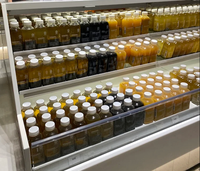 Reihen von Saftflaschen mit weißen Verschlüssen sind in einer Kühlvitrine in einem Geschäft fein säuberlich auf Regalen angeordnet. Die Flaschen enthalten verschiedene Saftfarben, darunter orange, gelb und dunkelviolett-schwarz.