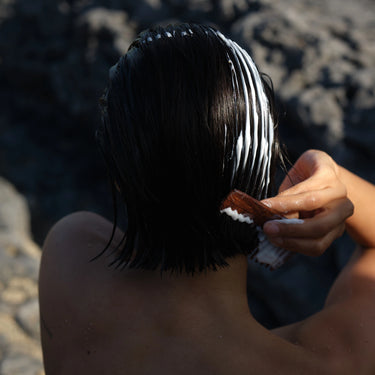 Eine Person mit nassem, dunklem Haar trägt DALUMA CLEAN HAIR CONDITIONER auf und kämmt es mit einem braunen Kamm im Freien in der Nähe von felsigem Terrain.