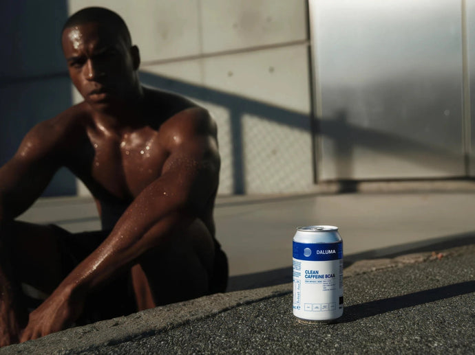 Ein Mann sitzt schwitzend und ohne Hemd auf dem Boden, im Hintergrund unscharf. Im Vordergrund steht eine Dose mit der Aufschrift "Clean Caffeine Dual" auf dem Bürgersteig, die scharf eingestellt ist.