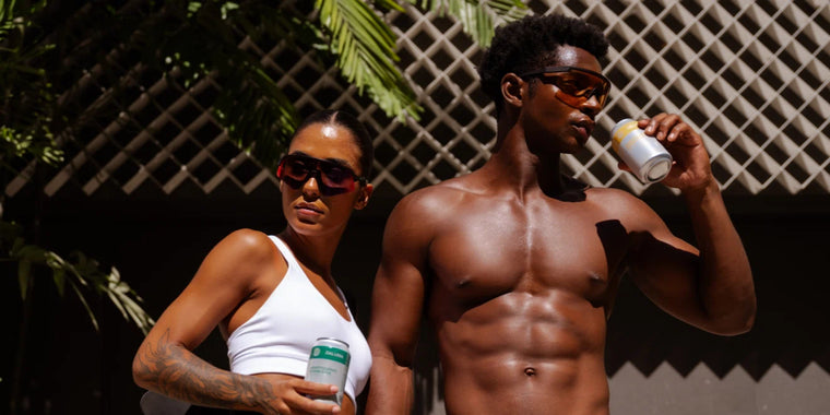 Zwei durchtrainierte Personen mit Sonnenbrillen und Sommerkleidung stehen im Freien im Sonnenlicht und halten jeweils ein Dosengetränk in der Hand; im Hintergrund sind Palmenwedel und ein Gitterzaun zu sehen.