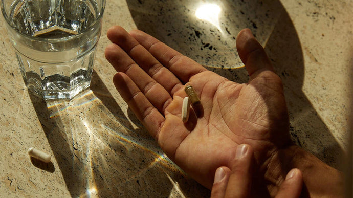 Eine Hand hält drei Kapseln oder Pillen über einer gesprenkelten Arbeitsplatte, daneben steht ein Glas Wasser und das Licht wirft Lichtreflexe auf die Oberfläche.