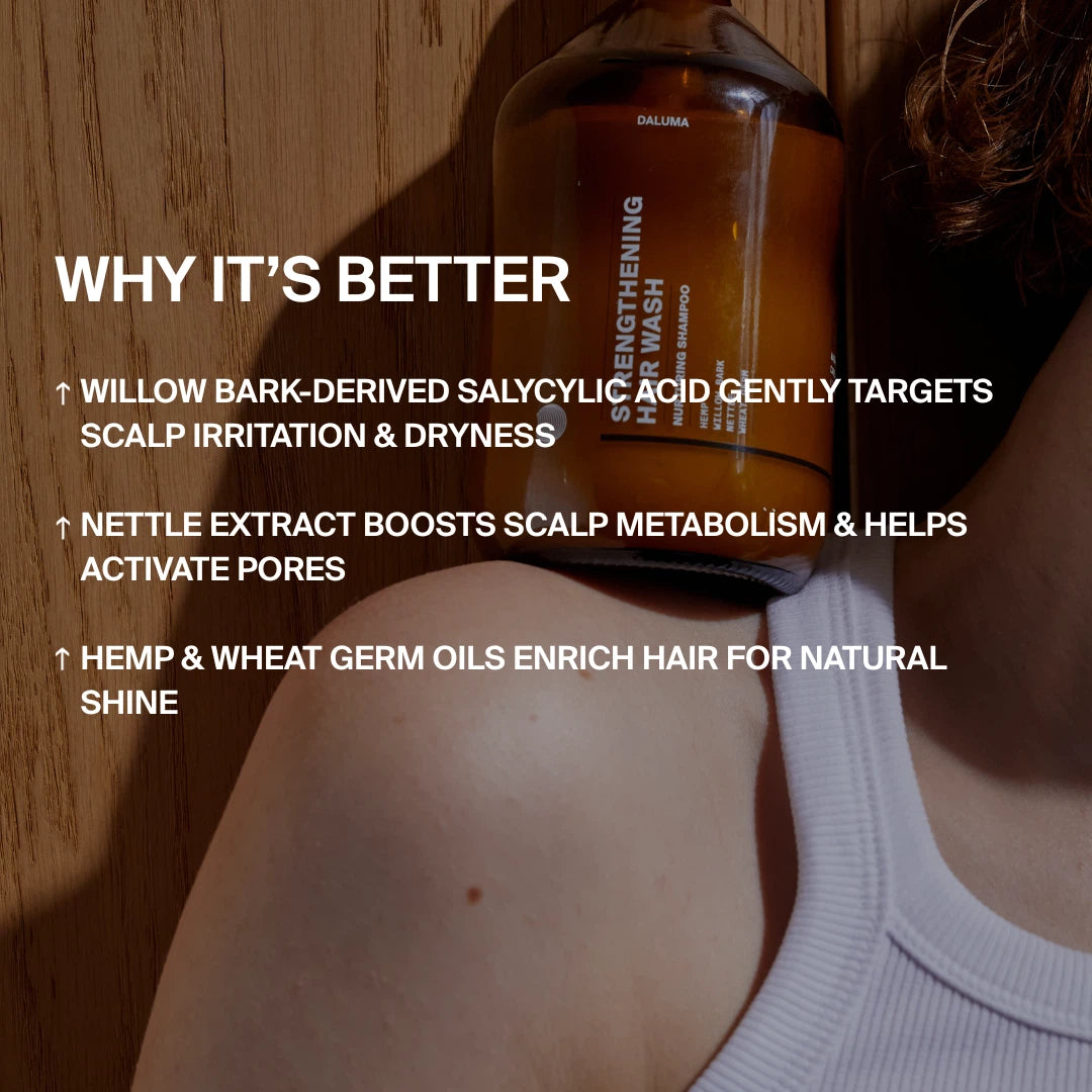 Eine braune Flasche mit der Aufschrift STRENGTHENING HAIR WASH von DALUMA ruht auf der Schulter einer Person. Der Text hebt natürliche Inhaltsstoffe hervor, die Kopfhautreizungen lindern, den Stoffwechsel anregen und dem Haar Glanz und Feuchtigkeit verleihen.