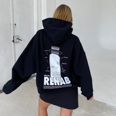 Eine Person mit langen blonden Haaren trägt den DALUMA JOSEPH ATELIER X DALUMA REHAB HOODIE mit grafischem Muster und dem Schriftzug "REHAB" auf dem Rücken, kombiniert mit einem schwarzen Rock und weißen Turnschuhen in einem minimalistischen weißen Raum.