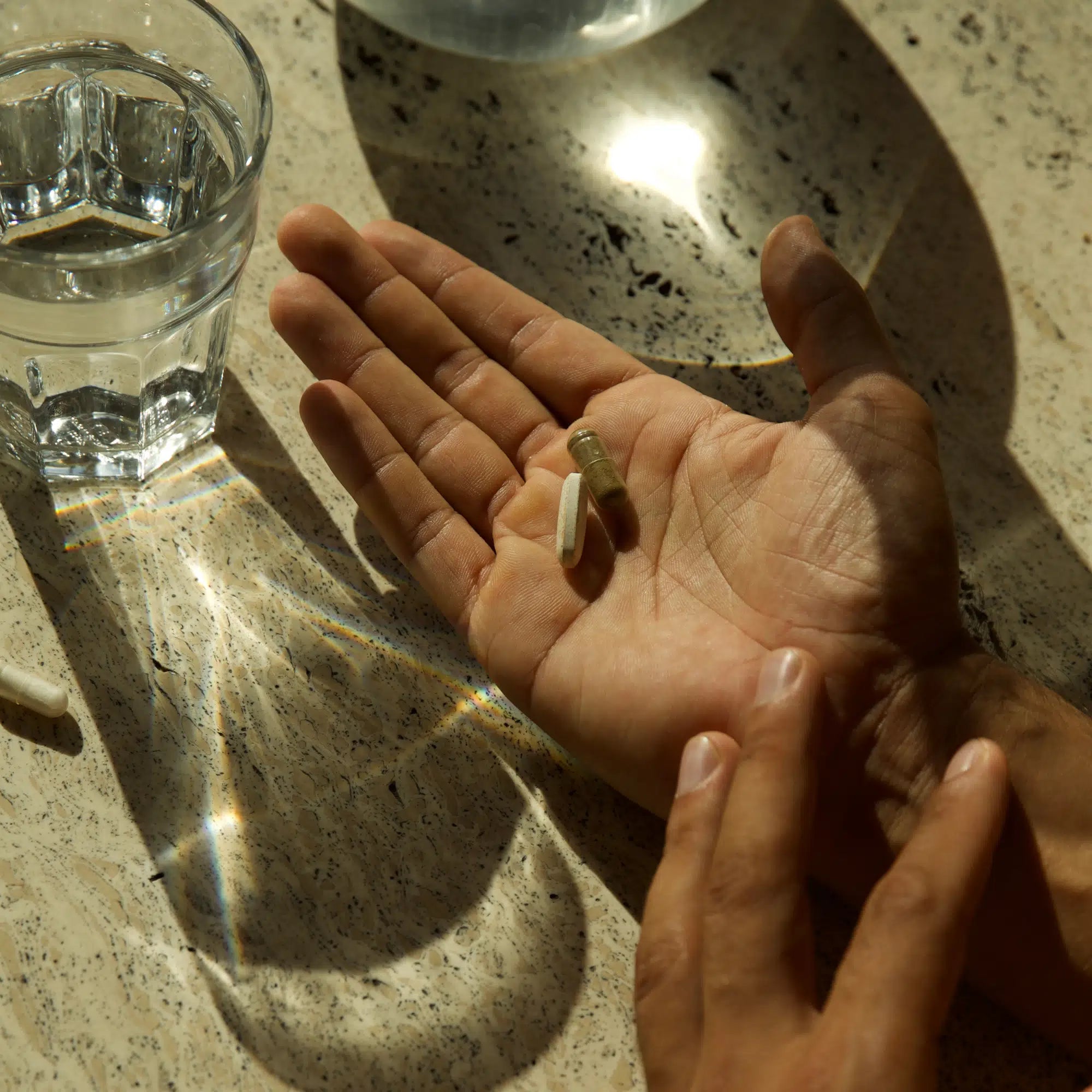 Eine Hand hält drei IMMUNE DEFENCE COMPLETE KOMPLEX DOUBLE Kapseln von DALUMA, während eine andere Hand nach ihnen greift; in der Nähe steht ein Glas Wasser auf einer sonnenbeschienenen, gesprenkelten Arbeitsplatte, das durch die Lichtreflexe auf der Oberfläche an die Unterstützung des Immunsystems erinnert.