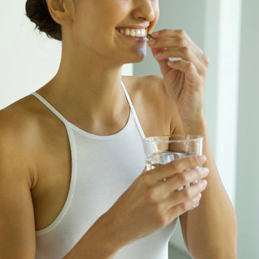 Eine Frau in einem weißen Tank-Top lächelt, während sie ein Glas Wasser in der Hand hält und sich auf die Einnahme einer DALUMA DEFENCE DUO Pille vorbereitet, die ihre Immunabwehr mit jeder Dosis unterstützt.
