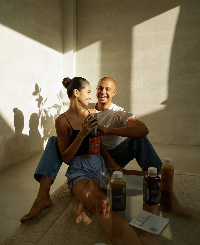 Ein lächelndes Paar sitzt auf dem Boden an einer sonnenbeschienenen Wand und hält Flaschen mit Saft in der Hand. Sie sind entspannt und glücklich. Auf einem Glastisch vor ihnen stehen weitere Getränkeflaschen und -pakete.