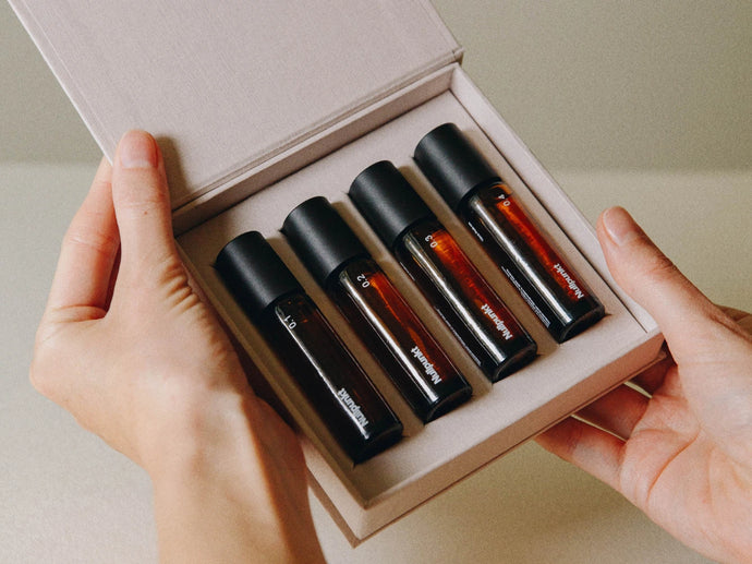 Die Hände halten eine offene Schachtel mit vier Braunglasflaschen mit schwarzen Verschlüssen, die jeweils mit weißem Text beschriftet sind. Die Flaschen sind ordentlich in einem hellen, gepolsterten Etui angeordnet.