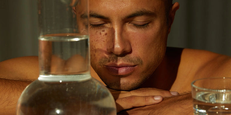 Ein Mann mit geschlossenen Augen stützt seinen Kopf auf seine verschränkten Arme auf einem Tisch. Sonnenlicht und Schatten fallen auf sein Gesicht. Im Vordergrund stehen ein Glas und eine klare Karaffe mit Wasser, die sein Gesicht teilweise verdecken.