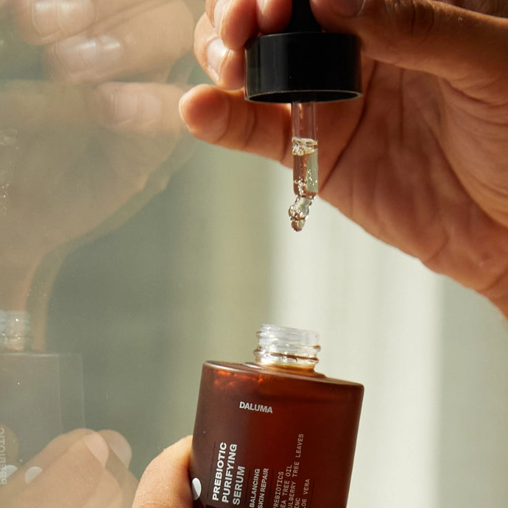 Eine Hand hält einen Tropfer mit klarem Serum über einer offenen braunen Flasche mit der Aufschrift DALUMA PREBIOTIC PURIFYING GESICHTSSERUM, während eine andere Hand die Flasche stützt.