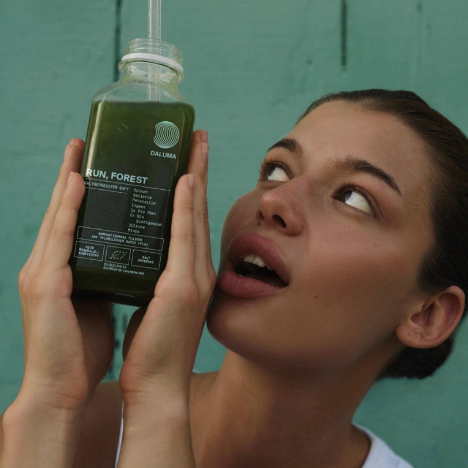 Eine Frau hält aufgeregt eine Flasche DALUMA RUN, FOREST GREEN JUICE mit einem Strohhalm hoch. Der kaltgepresste Saft ist mit dem Produktnamen beschriftet und hebt sich von einem einfarbig blaugrünen Hintergrund ab.
