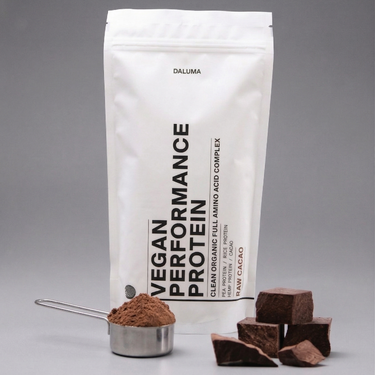 Ein weißer Beutel mit der Aufschrift "VEGANES PERFORMANCE PROTEIN VANILLE" von DALUMA steht aufrecht da. Davor ruht auf einer grauen Fläche ein Metalllöffel mit braunem Proteinpulver und rohen Kakaostückchen.