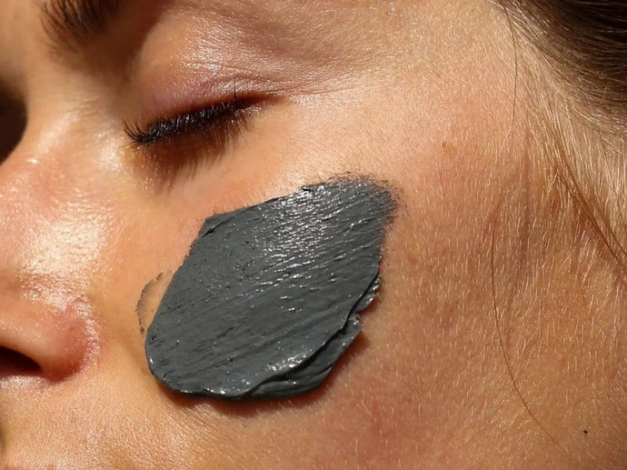 Eine Nahaufnahme des Gesichts einer Person mit geschlossenen Augen, die eine dicke Schicht einer grauen Tonmaske auf der Wange trägt. Die Haut wirkt glatt und natürlich.