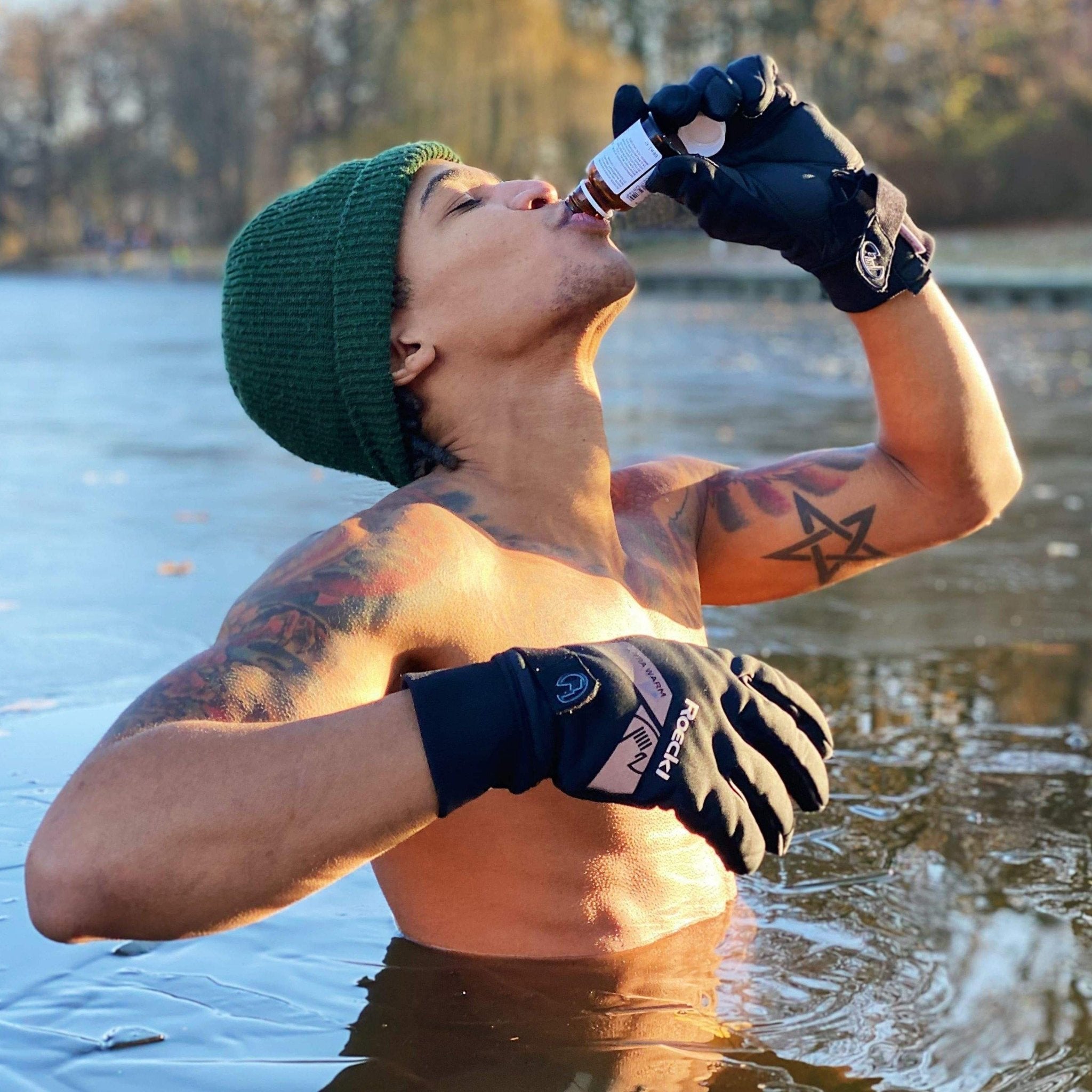 Eine Person ohne Hemd, mit grüner Mütze und Handschuhen steht in eisigem Wasser und trinkt einen DALUMA BURNING MAN INGWERSHOT 12 PACK, mit Bäumen und einer gefrorenen Landschaft im Hintergrund.