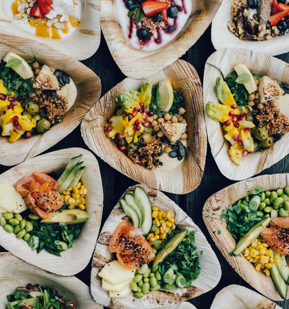 Verschiedene gesunde Salate und Schalen mit Gemüse, Getreide, Früchten, Räucherlachs, Samen und Grünzeug auf dreieckigen Holztellern auf einer dunklen Oberfläche.