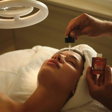 Eine Person entspannt sich mit einem in ein Handtuch gewickelten Kopf, während eine andere Hand das DALUMA CELLULAR RESURGENCE ANTI AGING SERUM, das kurzkettige Hyaluronsäure enthält, mit einer Pipette auf ihr Gesicht aufträgt - ein Hinweis auf eine feuchtigkeitsspendende Spa-Gesichtsbehandlung.