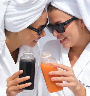 Zwei Frauen in weißen Bademänteln und mit Handtüchern auf dem Kopf lächeln und stoßen mit Sonnenbrillen auf Glasflaschen mit Saft an, eine dunkle und eine orange.