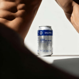 Eine Dose DALUMA CLEAN BCAA ENERGY DRINK 24 PACK liegt auf einer weißen Fläche, während eine unscharfe, muskulöse, hemdsärmelige Person im hell erleuchteten Vordergrund mit markanten Schatten steht.
