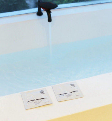 Eine Badewanne füllt sich mit Wasser aus einem schwarzen Wasserhahn. Im Vordergrund liegen zwei weiße DALUMA CLEANSING AKTIVKOHLE FACE MASK-Päckchen auf dem Wannenrand. Beide enthalten Bambuskohle und marokkanische Lavaerde.