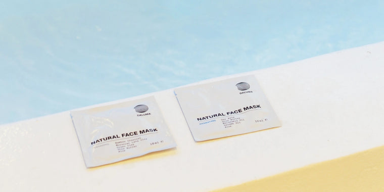 Zwei Pakete mit natürlichen Gesichtsmasken liegen nebeneinander auf einer weißen Leiste neben einem blauen Wasserbecken. Die Verpackung ist minimalistisch, mit schwarzem Text und einem kleinen runden Logo.