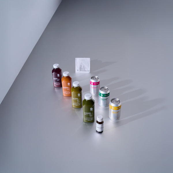 Verschiedene abgefüllte Säfte, Dosen, eine kleine Flasche und ein Tütchen sind ordentlich auf einer hellgrauen Fläche angeordnet und werfen weiche Schatten. Die Gegenstände sind in einem halbkreisförmigen Muster angeordnet.