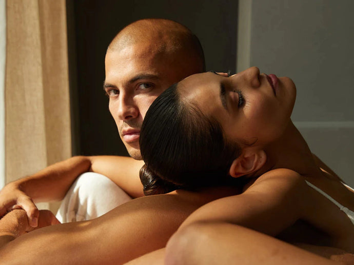 Ein Mann mit rasiertem Kopf blickt direkt in die Kamera, während eine Frau mit dunklem Haar sich an ihn lehnt und ihren Kopf auf seine Schulter stützt. Warmes Sonnenlicht beleuchtet ihre Gesichter und Oberkörper.
