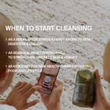 Jemand hält eine DALUMA BESSERE SAFTKUR PLUS - 7 TAGE grüne Saftflasche in der Hand, während er eine Cleanse-App benutzt. Das Bild zeigt den idealen Zeitpunkt für eine Saftkur: nach Stress, übermäßigem Essen, saisonal oder um gesunde Gewohnheiten zu beginnen. Ein leichter Teppich ist sichtbar.