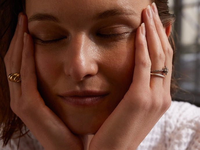 Eine Person mit geschlossenen Augen stützt ihr Gesicht in ihre Hände, trägt goldene und silberne Ringe und hat einen ruhigen und entspannten Ausdruck.