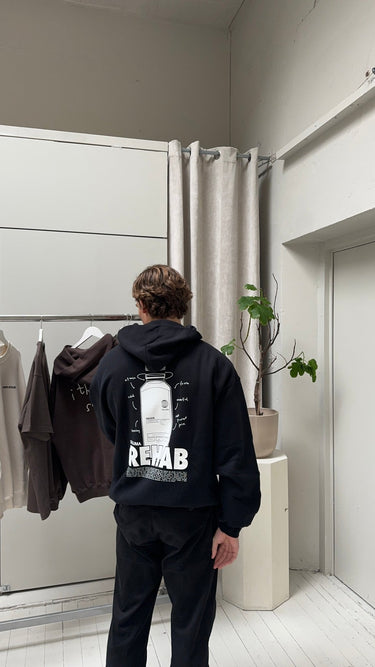 In einem minimalistischen Raum hält jemand in dem JOSEPH ATELIER X DALUMA REHAB HOODIE von DALUMA mit einer Grafik und "REHAB" auf dem Rücken einen braunen Kapuzenpullover aus Biobaumwolle von einem Ständer. Eine kleine Topfpflanze steht auf einem weißen Ständer in der Nähe.
