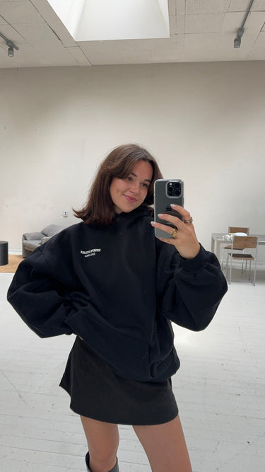 Eine Frau mit schulterlangen braunen Haaren trägt den JOSEPH ATELIER X DALUMA REHAB HOODIE von DALUMA und einen schwarzen Rock. Sie steht in einem minimalistischen, lichtdurchfluteten Raum und lächelt, während sie ein Selfie im Spiegel schießt - ein Zeichen für nachhaltige Mode.