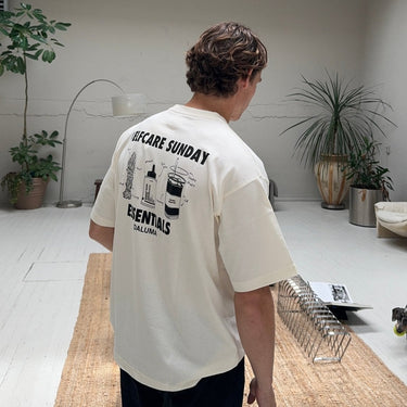Eine Person mit kurzen braunen Haaren steht in einem hellen, minimalistischen Raum mit Pflanzen, einem Teppich und einer Stehlampe. Sie trägt das DALUMA JOSEPH ATELIER X DALUMA SELFCARE SUNDAY TEE mit Oversize-Passform und illustrierter Rückengrafik.
